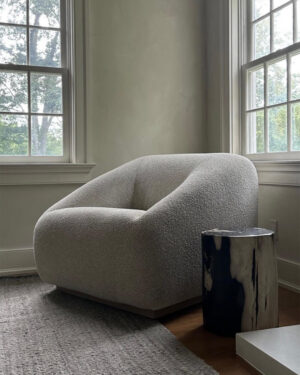 A curved armchair and sculptural wood side table sit near a window, placed over a smokey gray wool rug.