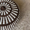 Detalle de almohadón redondo marrón chocolate y blanco natural con diseño peinecillo, sobre alfombra de lana.
