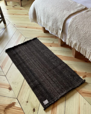 Close-up of a black handwoven wool rug placed on light wood flooring next to a bed with a beige throw blanke