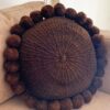 Chocolate brown-colored round cushion with handmade pom poms on a beige sofa.
