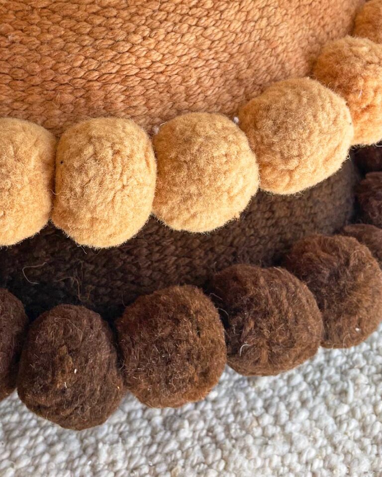 Almohadones con pompones en color camel y chocolate sobre una alfombra de lana de oveja de Andina Decor.