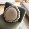 Almohadón redondo grande color blanco natural con pompones hechos a mano, acomodado sobre un sillón gris.