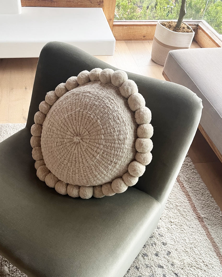 Natural pom pom cushion on a green sofa in a cozy living room by Andina Decor.