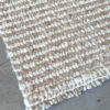 Esquina doblada de la Alfombra Rayada sobre un piso de cemento, mostrando su espesor y textura tejida a mano en beige (arena) y blanco (natural).