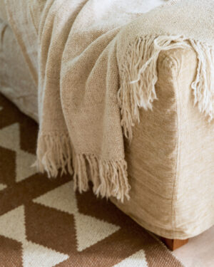 Light llama throw draped over the armrest of a beige sofa, with fringe details cascading down, next to a geometric patterned rug.