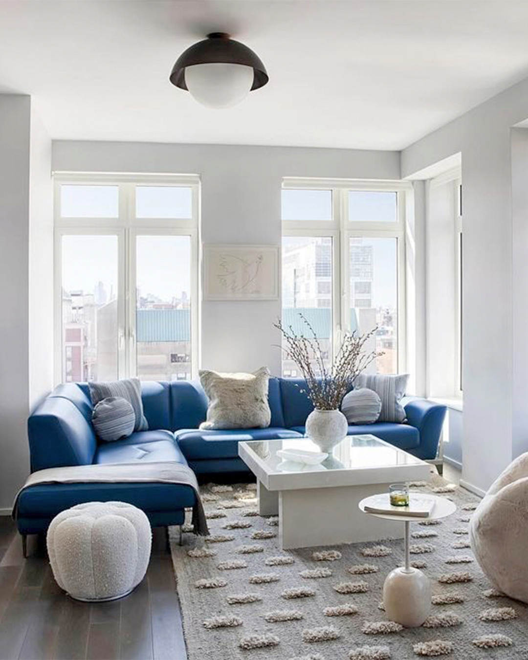 Alfombra gris humo con pompones blancos colocada bajo sofá azul y mesa baja blanca.