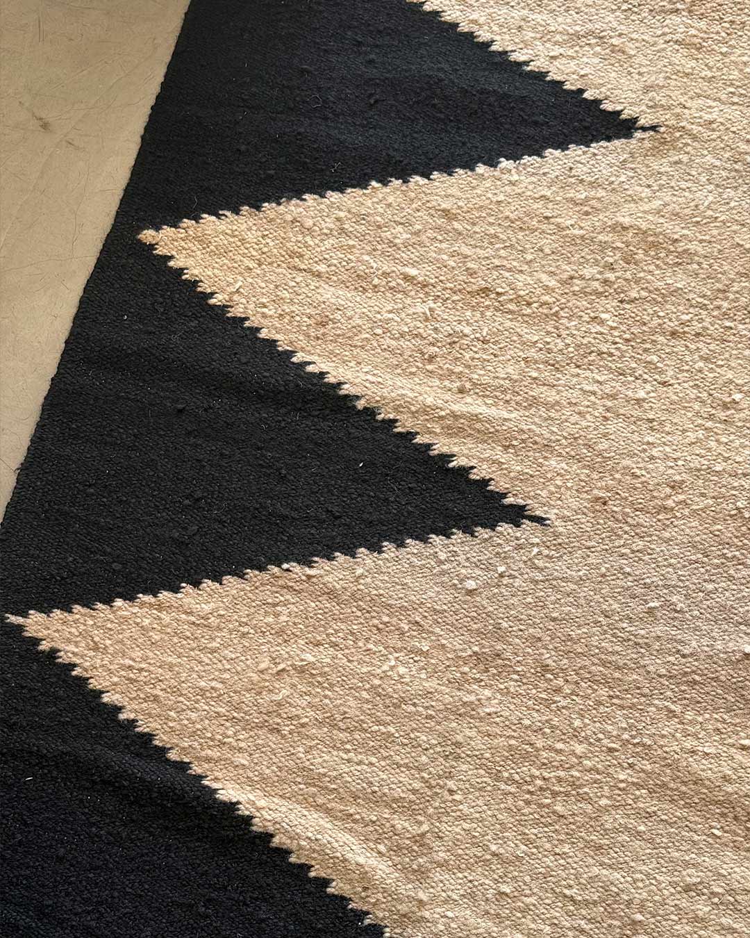 Vista en ángulo de la alfombra baetón tejida a mano con diseño diagonal en negro sobre base arena (beige).