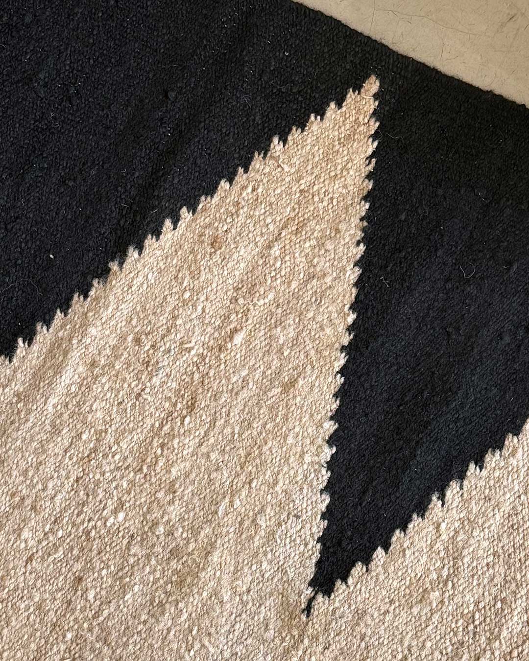 Detalle de una alfombra de lana tejida a mano con diseño geométrico en negro sobre base arena (beige).