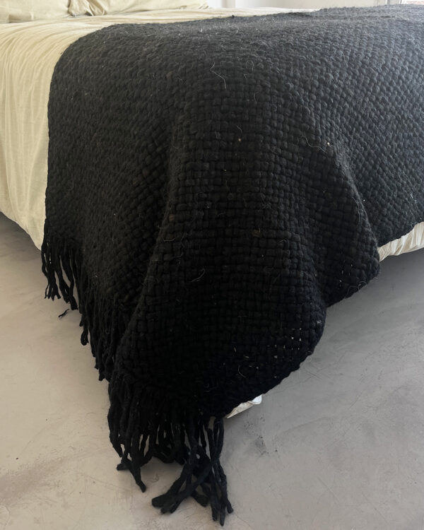 Handwoven black wool throw made from natural sheep's wool, with a thick texture and fringe detail, draped over a bed.