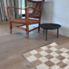 Wooden chair with a woven seat next to a small black table on light wood flooring, with a sand and natural checkered rug in the foreground.