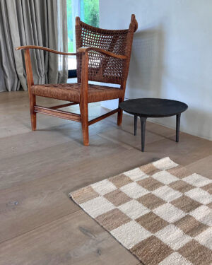 Wooden chair with a woven seat next to a small black table on light wood flooring, with a sand and natural checkered rug in the foreground.