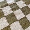 Close-up angled view of a wool rug with moss green and natural white checkerboard pattern, highlighting its rich woven texture.