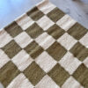 Handwoven wool runner rug with a checkered pattern in moss green and natural white, photographed at an angle on a polished concrete floor.