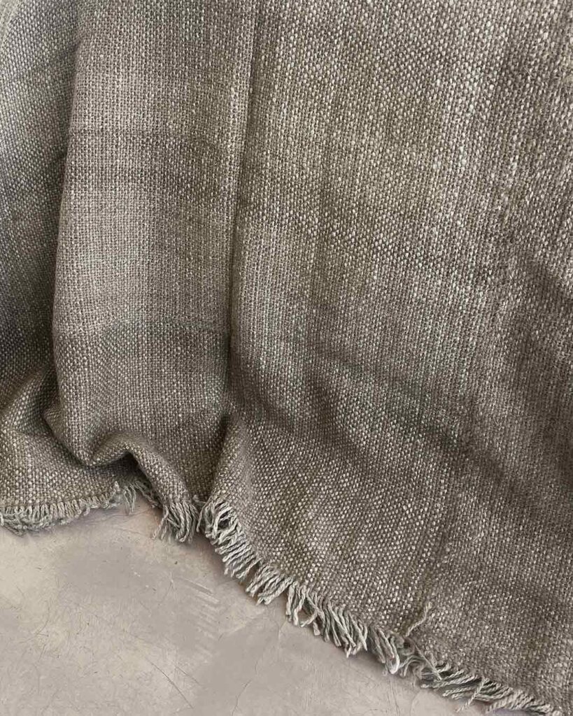 Llama bed quilt oat pale warm beige on concrete floor showing full handwoven structure and fringe